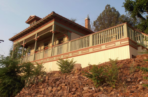 A colorful, two-story house with a porch, set on a hillside surrounded by greenery and rocks.